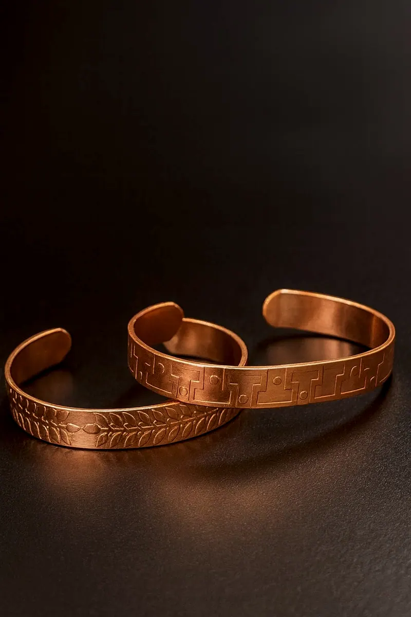 Duas pulseiras de cobre sobre fundo escuro: uma com folhas em relevo e outra com desenho geométrico em degraus e pontos